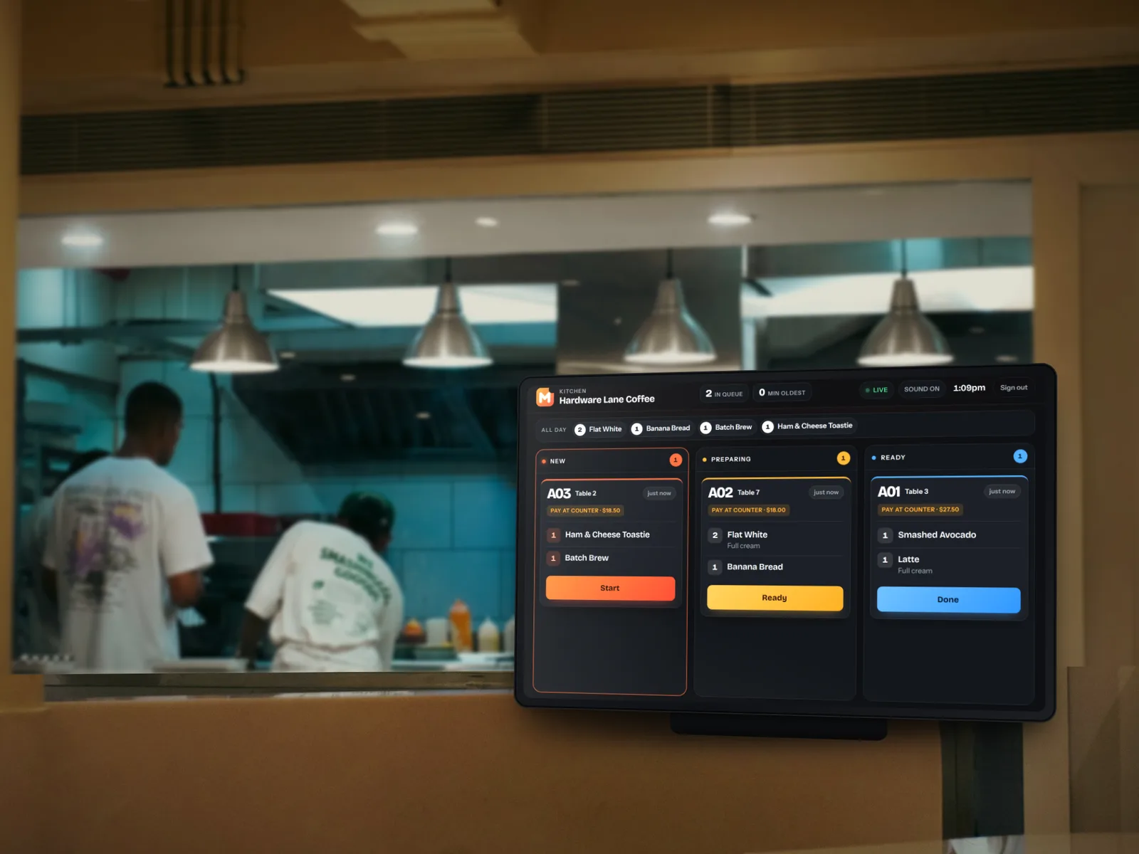 A kitchen screen on the pass showing the Munchly board: a new docket on the New rail, one preparing, one ready, with the kitchen at work behind