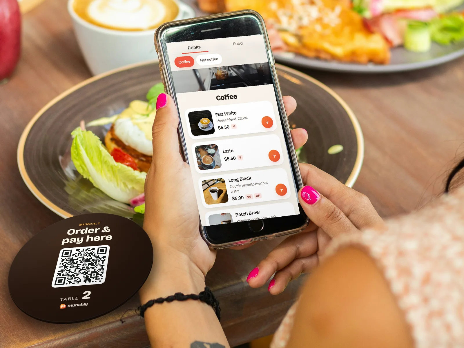 A hand holding a phone with the Hardware Lane Coffee menu open, over a cafe table with a round Munchly order-and-pay disc for Table 2 beside the plate
